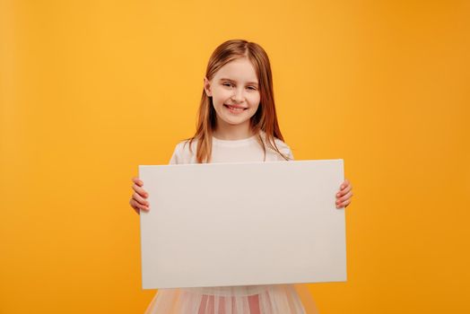 Pretty smiling girl child holding white canvas and looking at the camera isolated on yellow background with copyspace. Horizontal portrait of kid holding linen with place for text