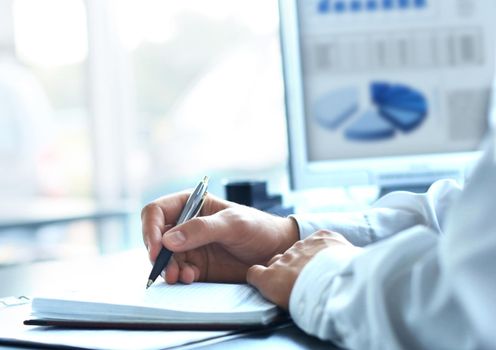 businessman writes in a notebook while sitting at a desk.