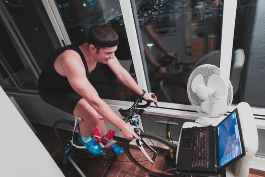 Man cycling on the machine trainer he is exercising in the home at night. Playing online bike racing game during coronavirus covid19 lockdown. New normal concept.