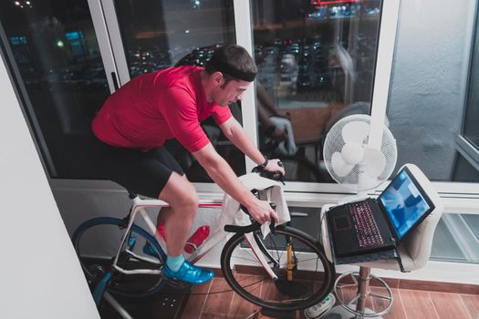 Man cycling on the machine trainer he is exercising in the home at night. Playing online bike racing game during coronavirus covid19 lockdown. New normal concept.