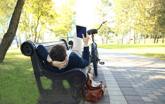 Businessman on a coffee break. He is sitting on a bench and working at touchpad, next to bike.