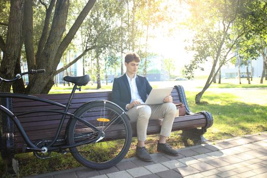 Businessman on a coffee break. He is sitting on a bench and working at laptop, next to bike.