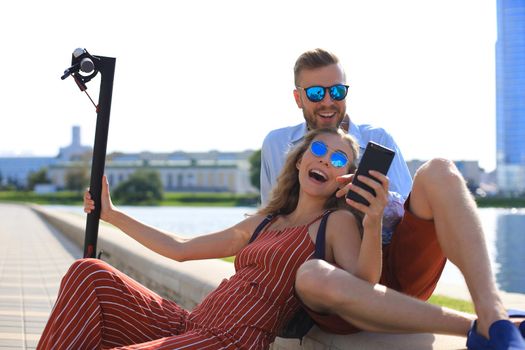 Lovely couple having fun driving electric scooter, taking a break from driving, sitting on riverbank, taking selfie,