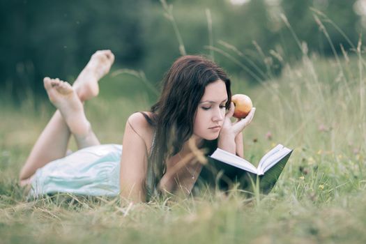 attentive girl with apple lying on the green grass and reading a book. the concept of a healthy lifestyle