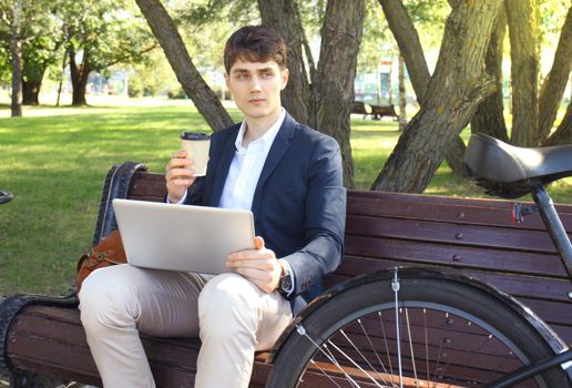 Businessman on a coffee break. He is sitting on a bench and working at laptop, next to bike.