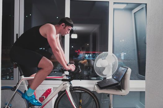 Man cycling on the machine trainer he is exercising in the home at night. Playing online bike racing game during coronavirus covid19 lockdown. New normal concept.