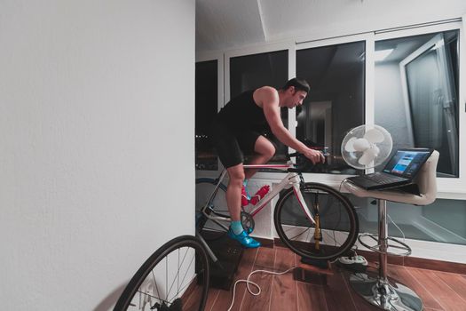 Man cycling on the machine trainer he is exercising in the home at night. Playing online bike racing game during coronavirus covid19 lockdown. New normal concept.