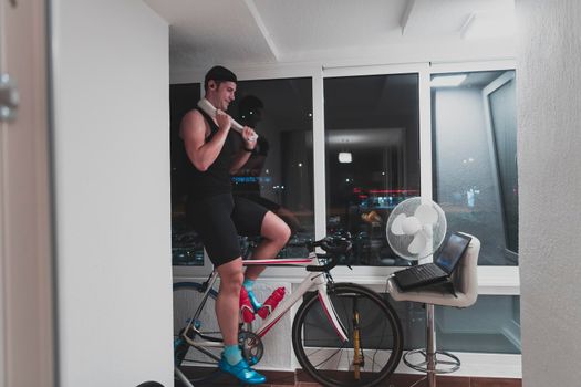 Man cycling on the machine trainer he is exercising in the home at night. Playing online bike racing game during coronavirus covid19 lockdown. New normal concept.