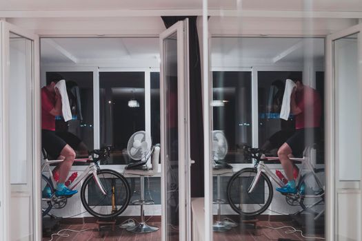 Man cycling on the machine trainer he is exercising in the home at night. Playing online bike racing game during coronavirus covid19 lockdown. New normal concept.