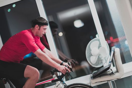 Man cycling on the machine trainer he is exercising in the home at night. Playing online bike racing game during coronavirus covid19 lockdown. New normal concept.