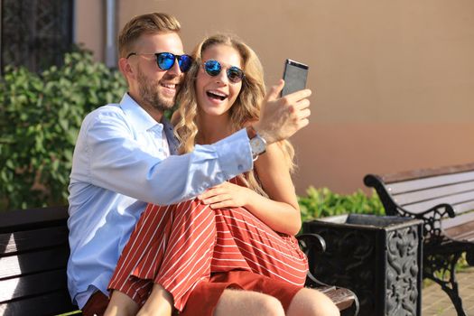 Romantic young couple in summer clothes smiling and taking selfie while sitting on bench in city street.