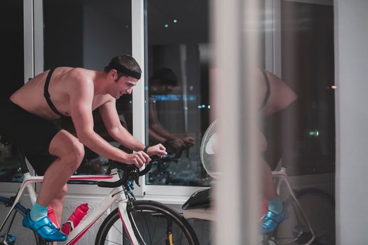 Man cycling on the machine trainer he is exercising in the home at night. Playing online bike racing game during coronavirus covid19 lockdown. New normal concept.