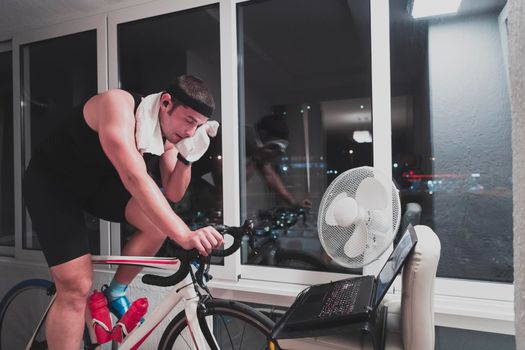 Man cycling on the machine trainer he is exercising in the home at night. Playing online bike racing game during coronavirus covid19 lockdown. New normal concept.