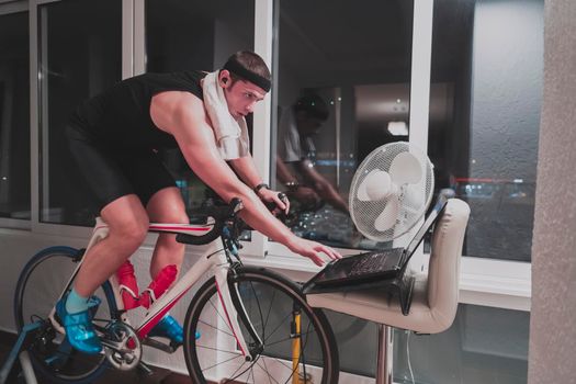 Man cycling on the machine trainer he is exercising in the home at night. Playing online bike racing game during coronavirus covid19 lockdown. New normal concept.