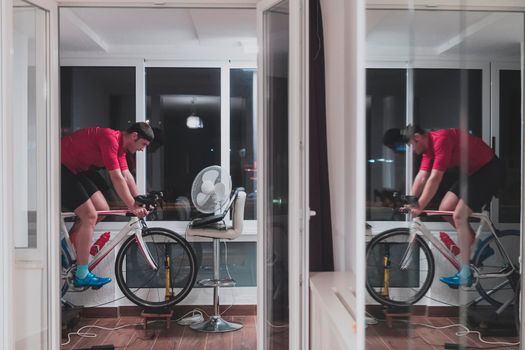 Man cycling on the machine trainer he is exercising in the home at night. Playing online bike racing game during coronavirus covid19 lockdown. New normal concept.