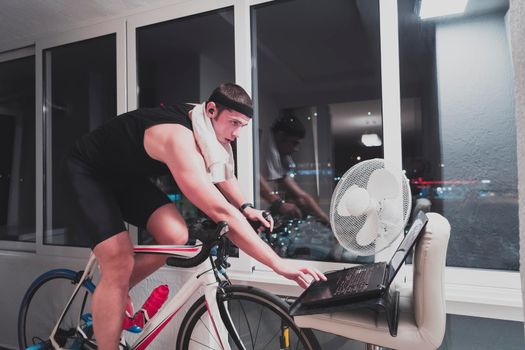 Man cycling on the machine trainer he is exercising in the home at night. Playing online bike racing game during coronavirus covid19 lockdown. New normal concept.