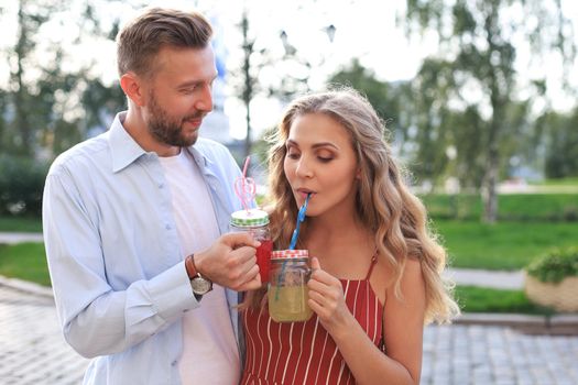 Beautiful young couple walking through the city street, smiling and drinking fresh cocktails.