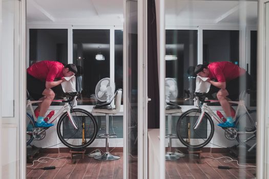 Man cycling on the machine trainer he is exercising in the home at night. Playing online bike racing game during coronavirus covid19 lockdown. New normal concept.