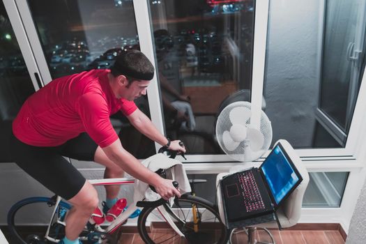 Man cycling on the machine trainer he is exercising in the home at night. Playing online bike racing game during coronavirus covid19 lockdown. New normal concept.