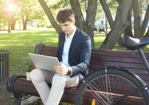 Businessman on a coffee break. He is sitting on a bench and working at touchpad, next to bike.