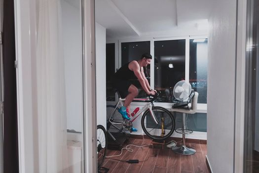 Man cycling on the machine trainer he is exercising in the home at night. Playing online bike racing game during coronavirus covid19 lockdown. New normal concept.