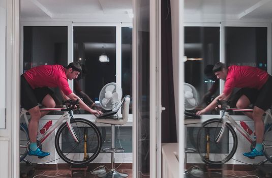 Man cycling on the machine trainer he is exercising in the home at night. Playing online bike racing game during coronavirus covid19 lockdown. New normal concept.