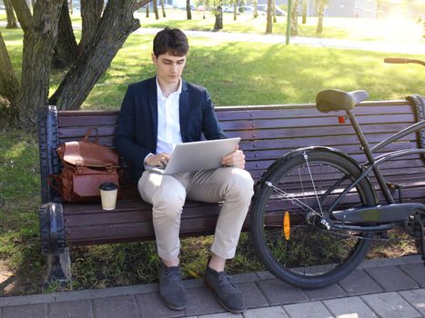 Businessman on a coffee break. He is sitting on a bench and working at laptop, next to bike.