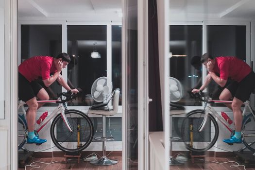 Man cycling on the machine trainer he is exercising in the home at night. Playing online bike racing game during coronavirus covid19 lockdown. New normal concept.