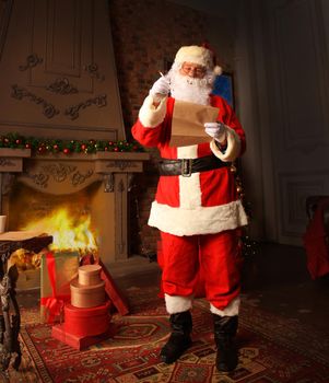 Santa Claus standing at his room at home near Christmas tree and big sack and reading Christmas letter or wish list.