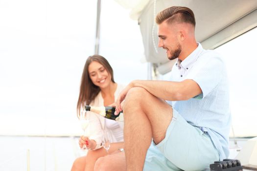 Smiling young couple with champagne and looking at each other while sitting on the board of yacht.