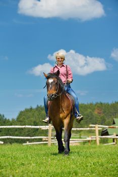 happy woman in sunglasses sitting on horse farm animal outdoors with blue sky in background