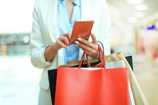 Woman using smartphone and holding shopping bag while standing on the mall background.