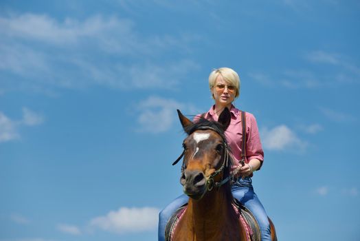 happy woman in sunglasses sitting on horse farm animal outdoors with blue sky in background