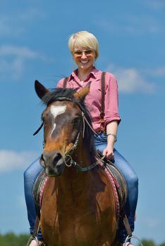 happy woman in sunglasses sitting on horse farm animal outdoors with blue sky in background