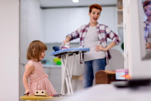 happy family having fun together at home  cute little daughter in a pink dress playing and dancing while young redhead mother ironing clothes behind her