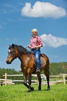 happy woman in sunglasses sitting on horse farm animal outdoors with blue sky in background