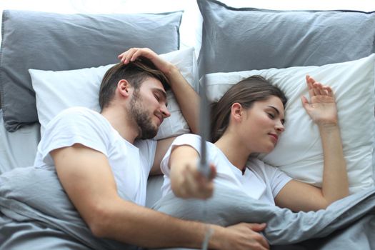 Beautiful young loving couple sleep and making selfie with stick in the bed.