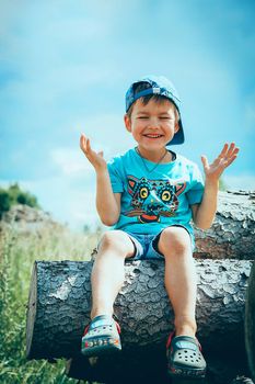 A preschool child in a baseball cap and denim shorts sits on a log playing and laughing