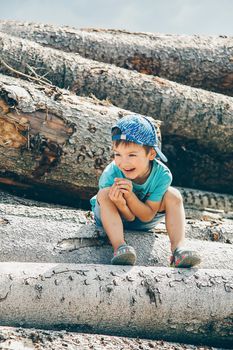 A little boy in a blue baseball cap and short shorts sits on a log and laughs merrily