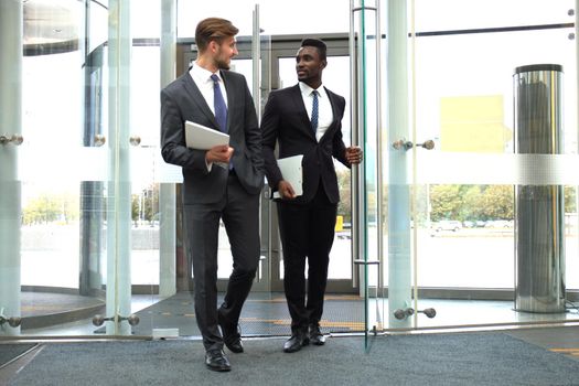 Two multinational young businessmen entering in office building with glass doors.