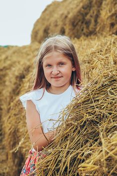 A blonde schoolgirl in a pink dress climbed on large bales of straw on a hot summer day