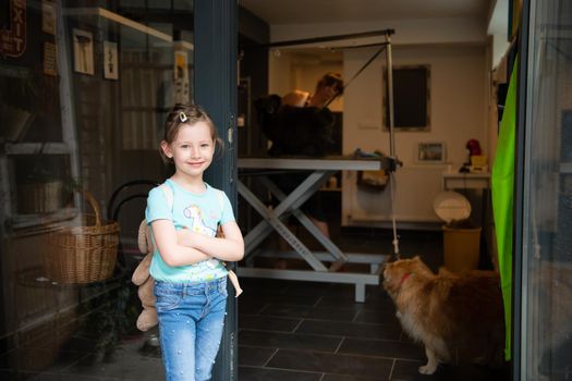 portrait of cute little girl standing and waiting while professional pet hairdresser finishing her black dog hairstyle in front of beauty salon for animals