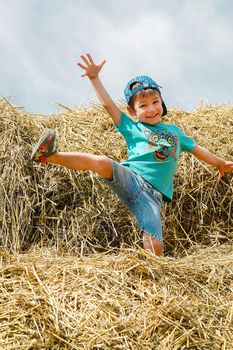 A schoolboy in a blue baseball cap and short shorts plays and has fun on straw bales on a hot summer day