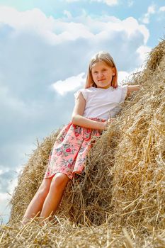 A beautiful blonde girl in a pink dress climbed on large bales of straw on a summer day