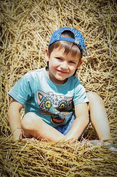 A boy in a blue baseball cap sits on a haystack and smiles on a hot summer day