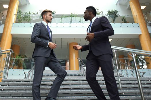 Two multinational young businessmen talking while stairs in modern office building.