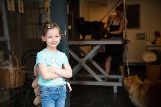 portrait of cute little girl standing and waiting while professional pet hairdresser finishing her black dog hairstyle in front of beauty salon for animals