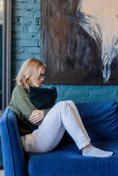 Young thoughtful caucasian woman in green sweater and white jeans sitting on sofa with eyes closed in stylish blue living room