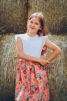 A blonde girl with long hair loose and in a dress stands near rolls of straw on a Sunny hot day