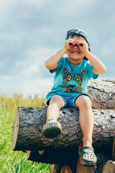 A schoolboy in a blue baseball cap and denim shorts sits on a log and peers into the distance